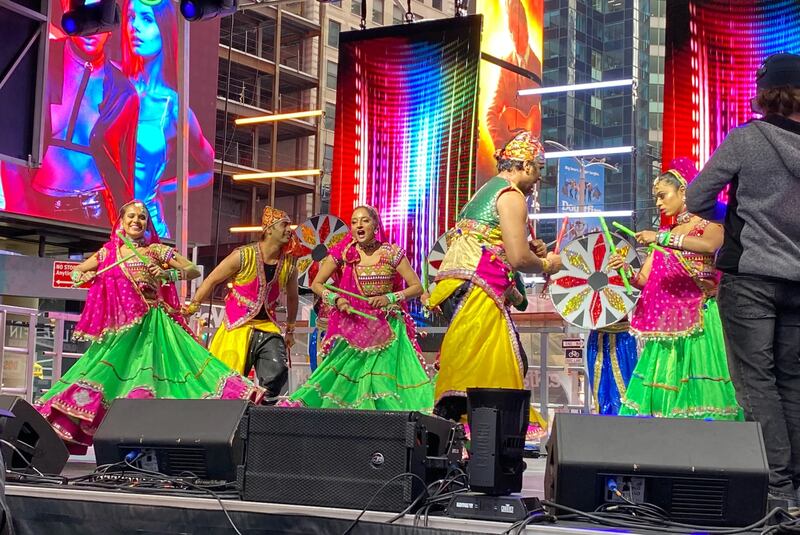 Men and women wearing bright pink, green and yellow traditional clothing dance and play percussion instruments on a stage in Manhattan’s Times Square.
