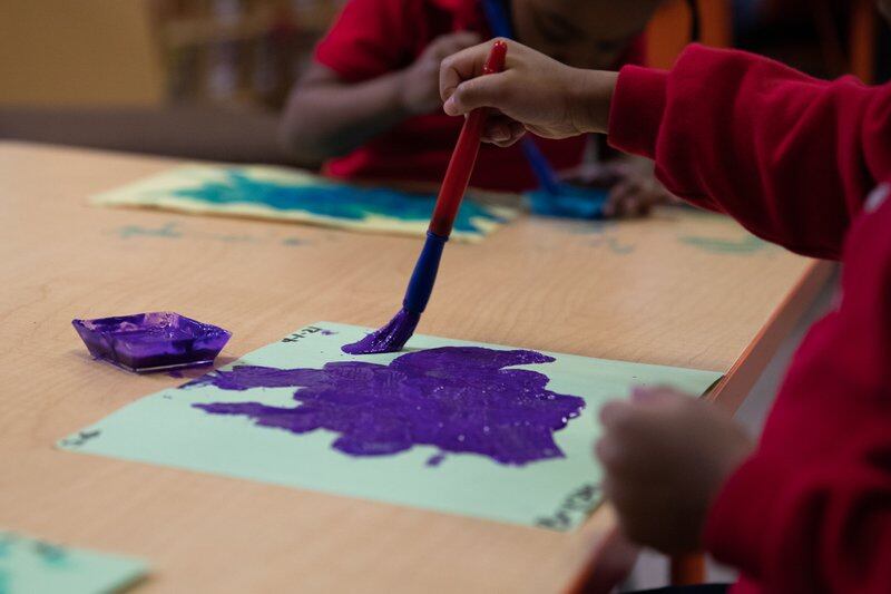 Two preschoolers paint with brushes in purple and turquoise on paper at a desk.