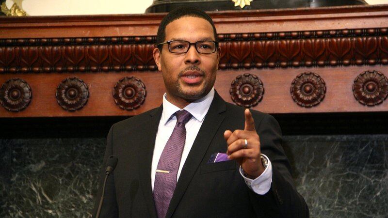 A man in a light shirt and dark suit with a purple pocket square and wearing glasses points his finger while giving a speech at a press conference.
