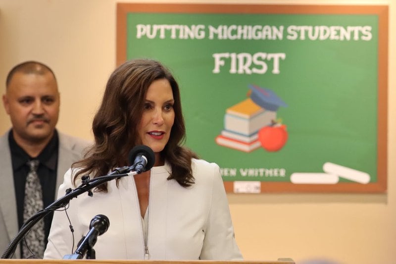 Michigan Governor Gretchen Whitmer speaks into a microphone in front of a sign that reads, “Putting Michigan Students First.” There is a man in a grey suit behind her right shoulder.