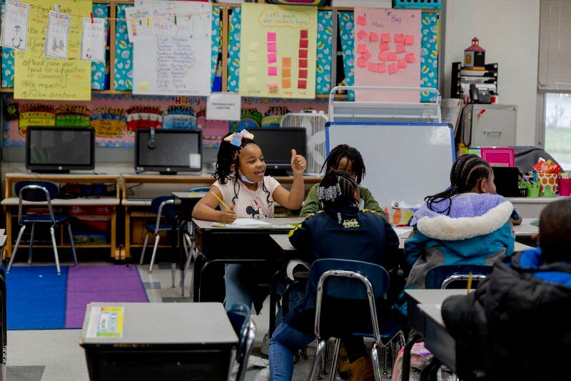 A young girl with a bow in her hair sits at a desk and talks with three other students in a classroom with colorful bulletin boards. 