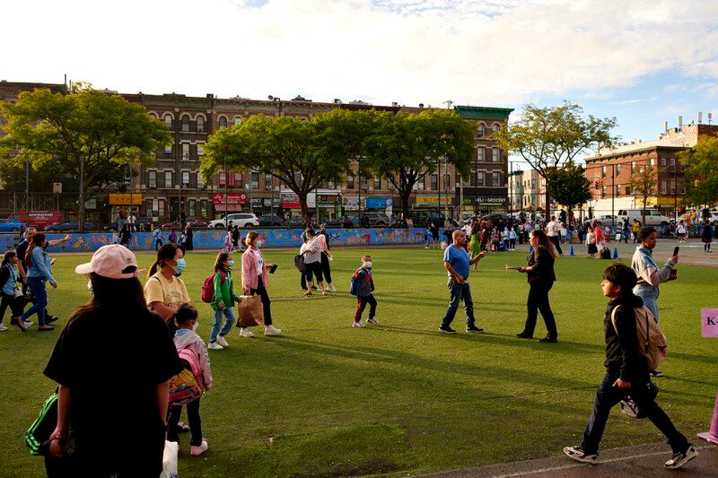 Parents and children, some in face masks, walk across an astroturf field with brick walkup apartment buildings in the background.