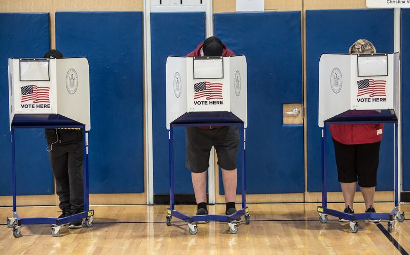 Three people stand behind voting partitions against a blue divider.