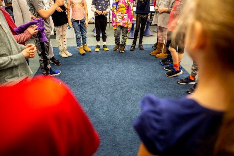 Students standing in a circle on a rug in a room, with their hands clasped, are shown from the waist down