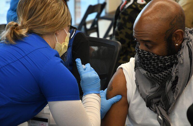 A woman wearing gloves and a mask gives a vaccine to a man wearing a mask.