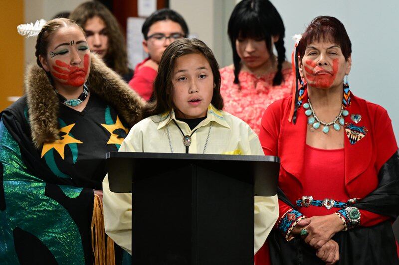A Native American student stands at a podium, addressing the school board. His mother and grandmother stand on either side of him. All are wearing traditional dress. His mother and grandmother have red painted handprints over their mouths.