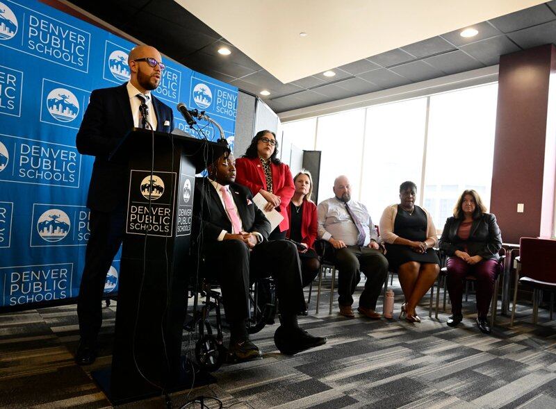 Denver Superintendent Alex Marrero, left, speaks at a podium. Denver school board members, including Board President Xóchitl “Sochi” Gaytán in a red blazer, are standing to the right. The Denver Public Schools logo is visible behind them.