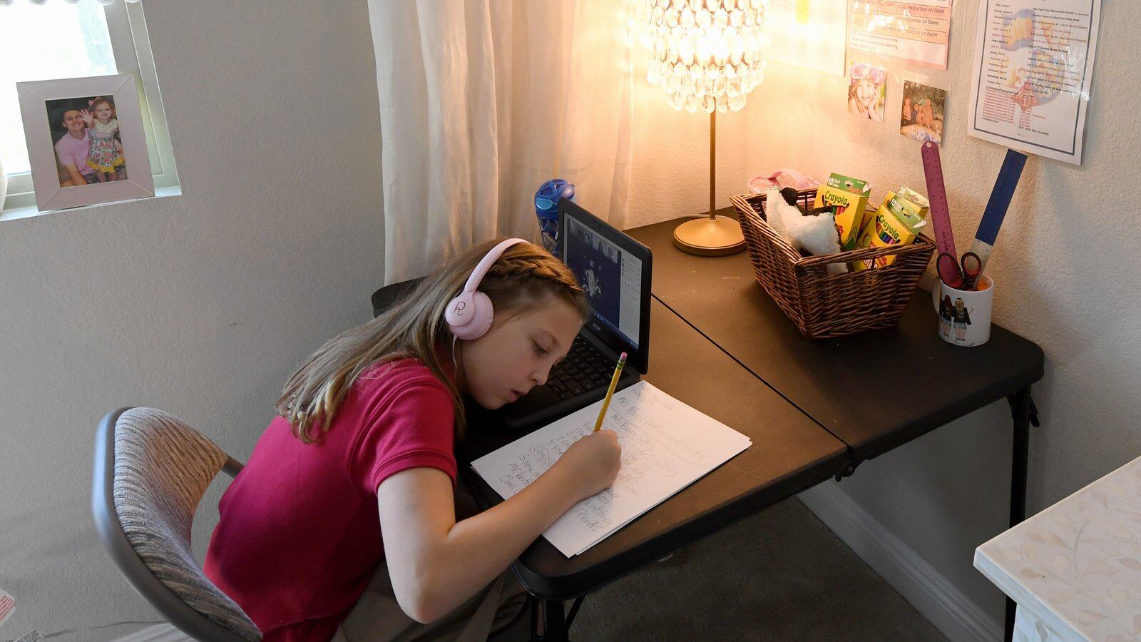 A young girl wearing a pink shirt and headphones writes on a piece of paper while sitting at her desk with a laptop open in front of her.
