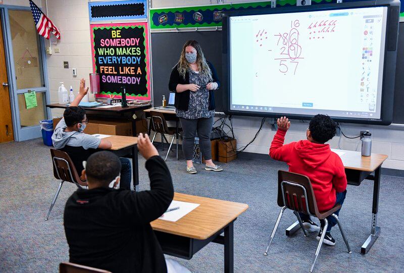 A teacher works through math problems on a smart board as her students raise their hands at their desks.