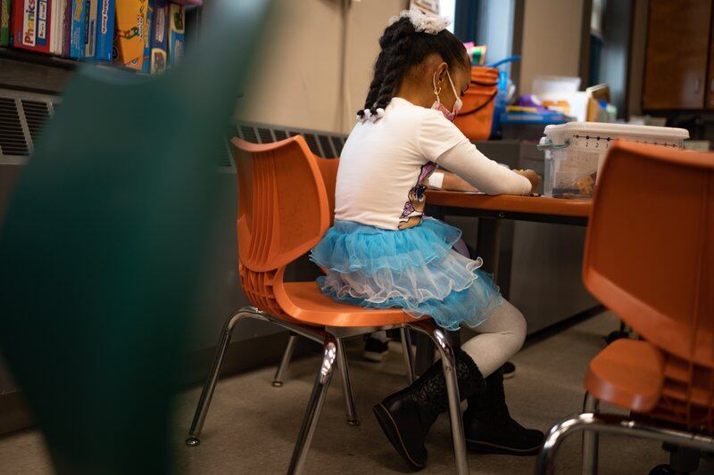 A young girl in a blue tutu skirt works at a desk, seen behind green and orange chairs.