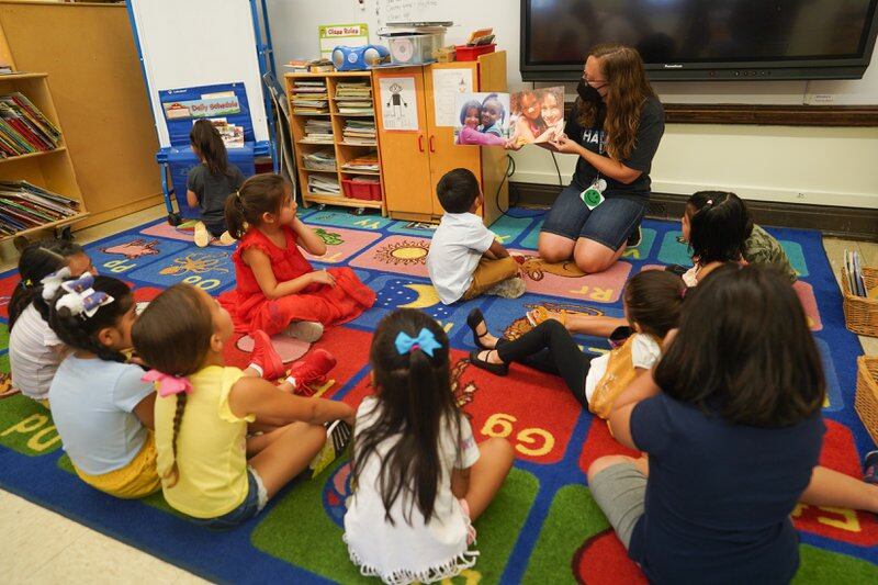 A teacher reads a book out loud in front of a group students who are sitting on colorful rug in a classroom.