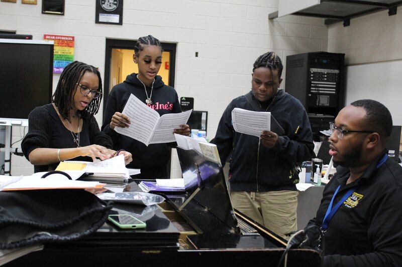 Three students stand on the side of a piano, singing while holding music sheets as their teacher plays the piano.