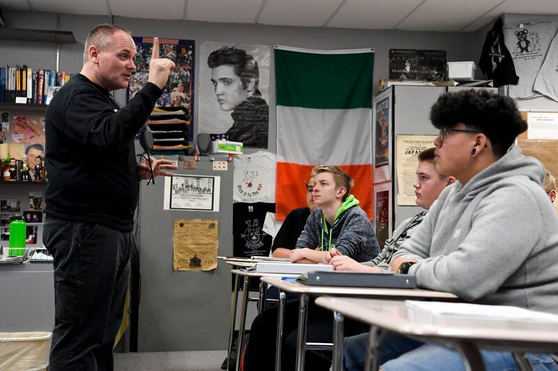 A male teacher stands in front of students at desks. A poster of Elvis Presley is on the wall behind them.