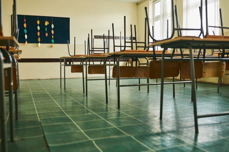 A classroom with a green tiled floor features rows of empty desks with chairs stacked on top of each desk.
