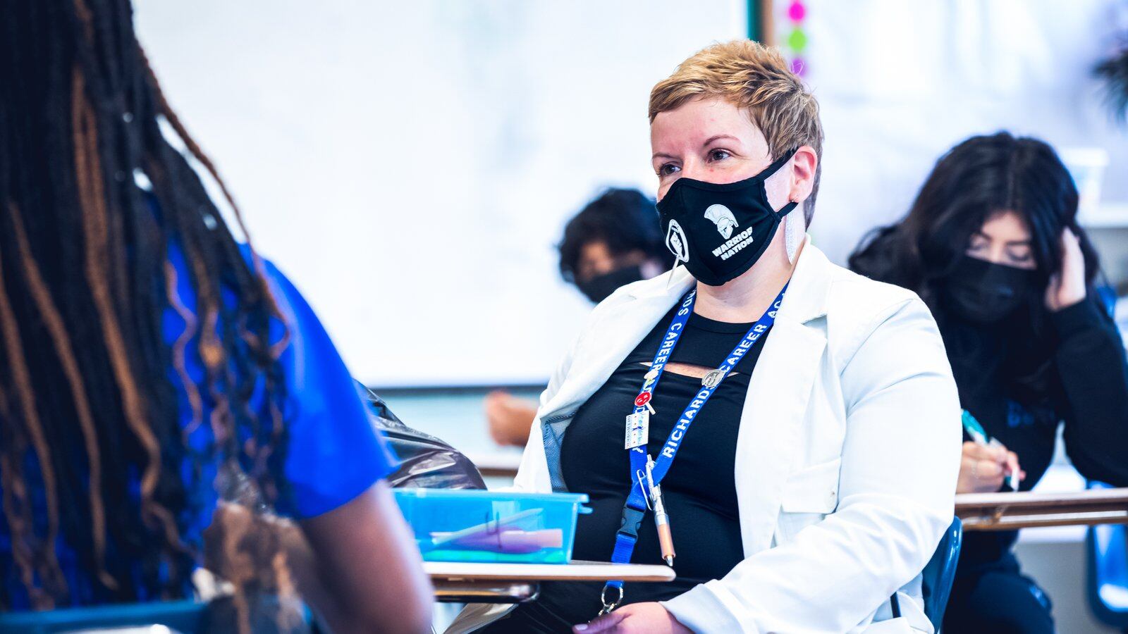 A woman in a face mask sits at a desk and speaks with a girl in a school classroom.