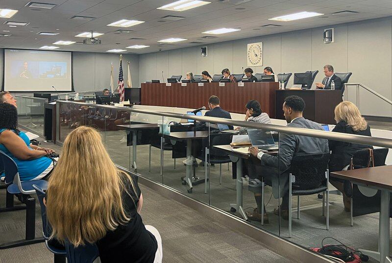 School district officials sit behind a glass barrier. School board members are behind them on an elevated platform.