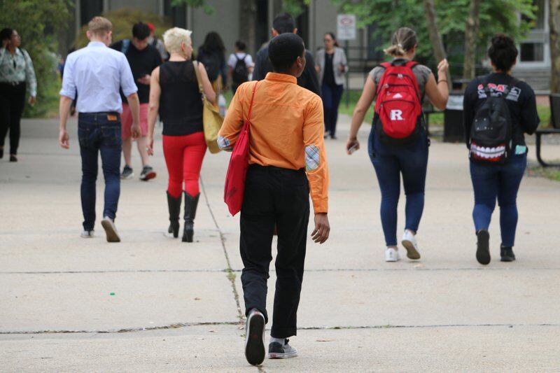 A young man wearing an orange shirt, black pants and carrying a red bag walks on a college campus behind other college students.