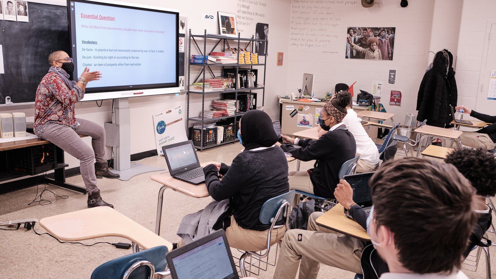 A teacher points at a screen while sitting on a desk in front of a group of students sitting at desks with laptops.  