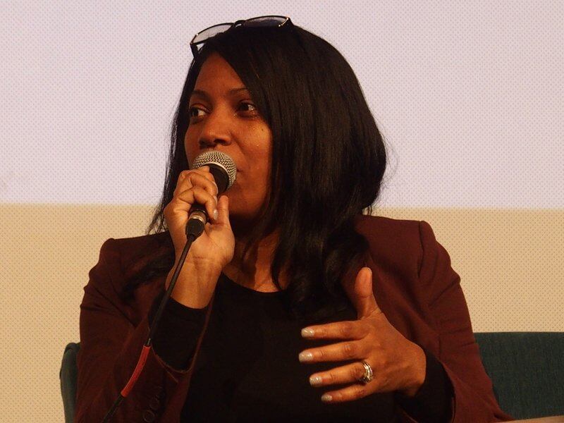 A woman with long dark hair and sunglasses on her head speaks into a microphone. She is wearing black clothes.