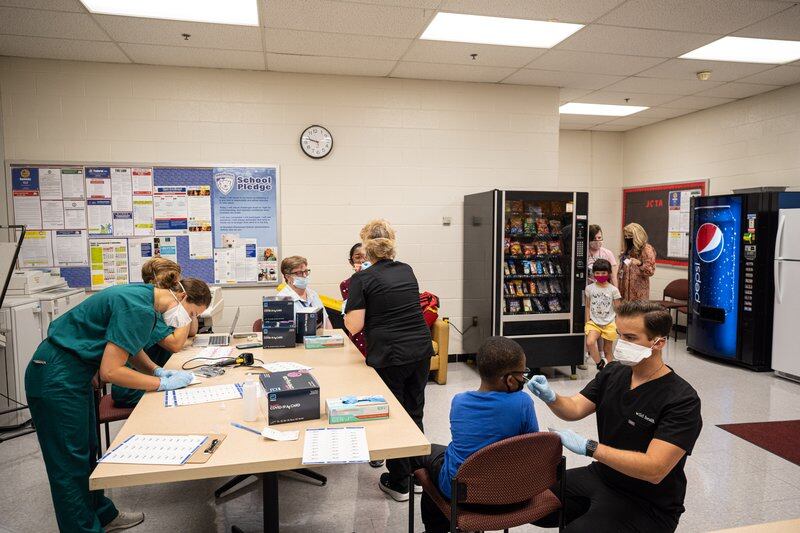 Medical professionals wearing face masks conduct COVID PCR testing in a room at a school.