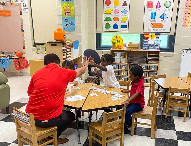 Three children sit around a table to work on an assignment in a colorful classroom. The educator, who is wearing a red shirt, gives a high five to a young student who is wearing a white collared shirt. 