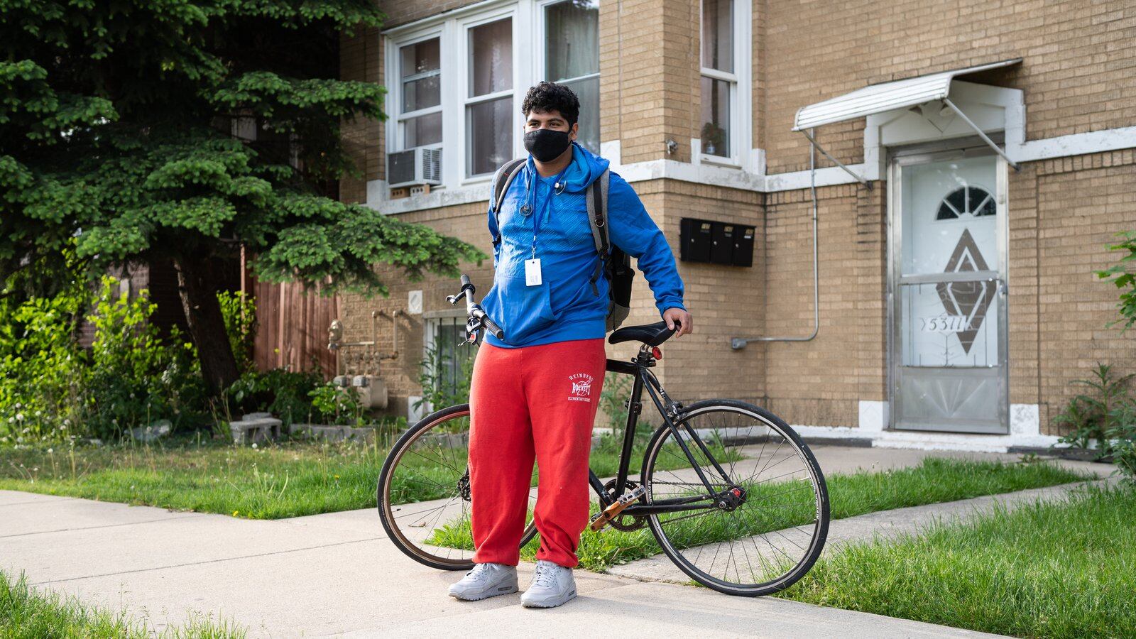 A young man, wearing a blue jacket, protective mask, and red track pants, poses for a portrait with his bike before riding to his high school.