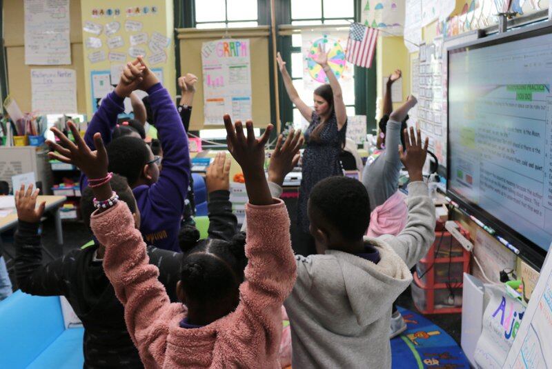 Students and a teacher raise their hands in a classroom.