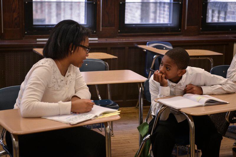 A student and a teacher sit at desks and talk to each other in a classroom while workbooks are open in front of them.