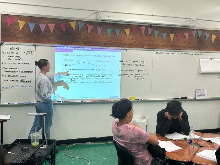 A teacher stands at a lit-up white board in a classroom. She is wearing a light shirt and jeans. Two students sit at their desks, studying. They both have dark hair.
