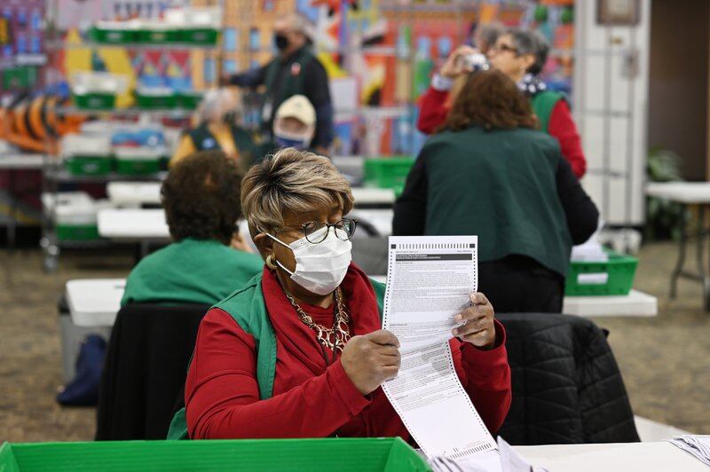 An election judge, wearing a protective mask and red jacket, sorts through ballots as other poll workers fill the background.