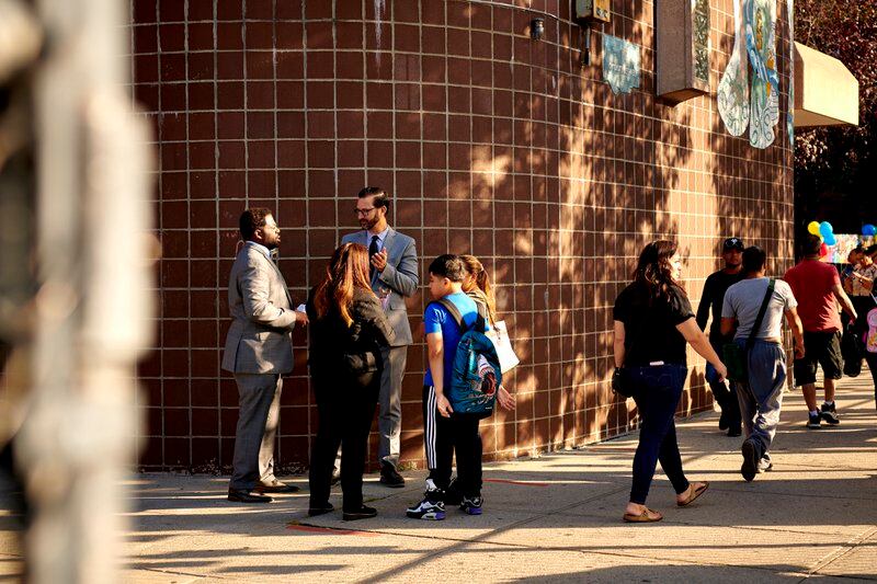 School administrators speak with a family on the first day of school as other parents walk by them outside a brick school building.