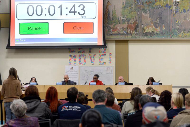 Denver school board members sit at a dais in a gymnasium. The audience sits in folding chairs with their backs to the camera. A big screen shows a stopwatch counting down.