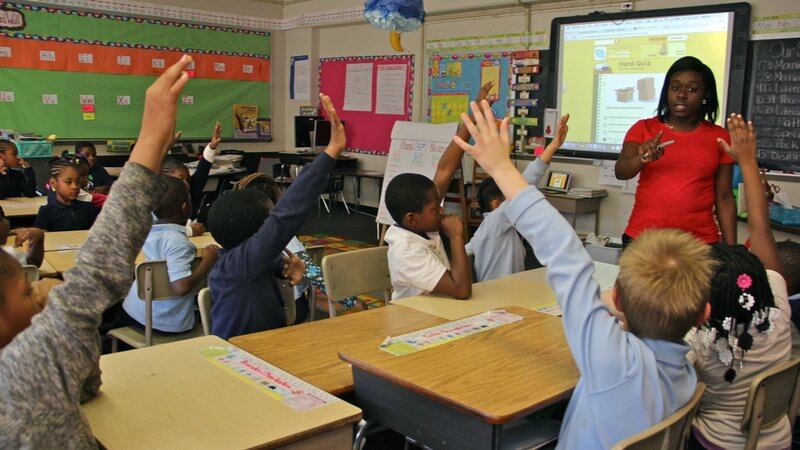 Students raise their hands at their desks while a teacher points to them at the front of the classroom.