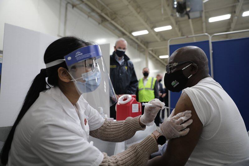 A healthcare worker wearing a face shield and mask gives a shot of a COVID vaccine to a man wearing a black mask and glasses.
