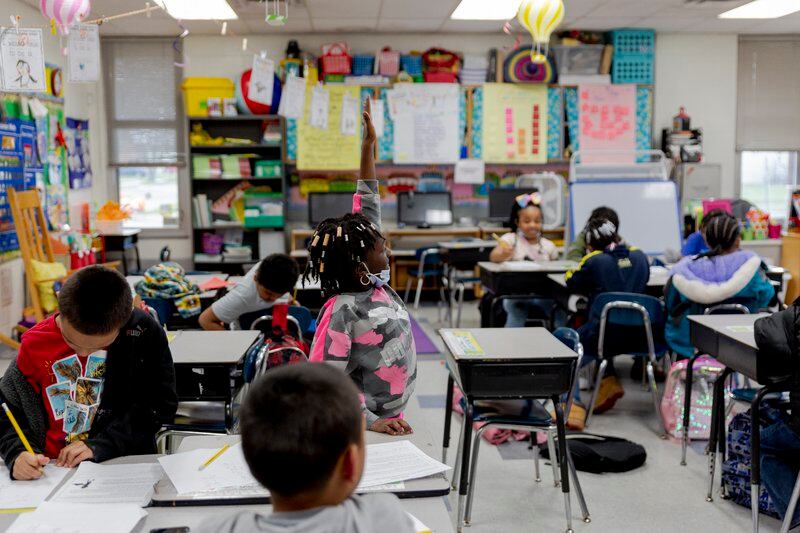 A student raises her hand and stands at her desk, as several other young students work around her in the classroom.