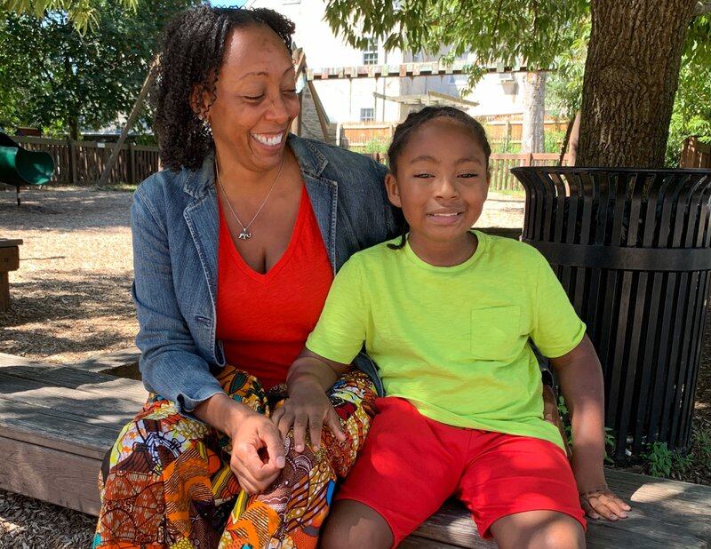 Jeannine Payne sits on a bench with her son Andre. She wears a red top and a denim jacket. He’s wearing a neon green shirt and red shorts.