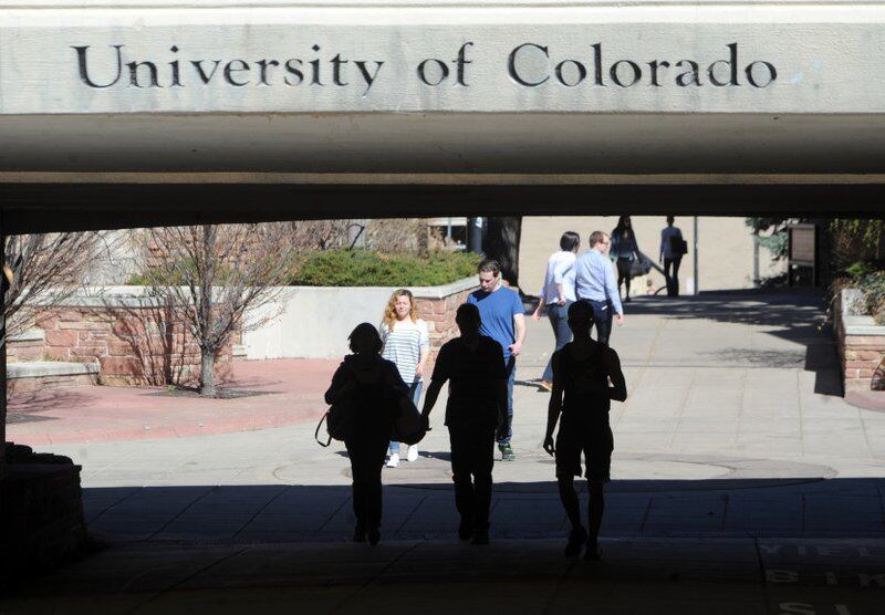 Several people walk underneath a grey bridge with the words “University of Colorado” etched into the concrete.