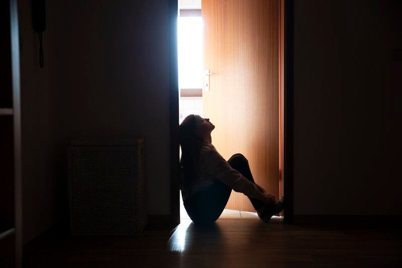A teenage girl sits in a door frame, silhouetted by light coming in through the door into a dark hallway behind her.