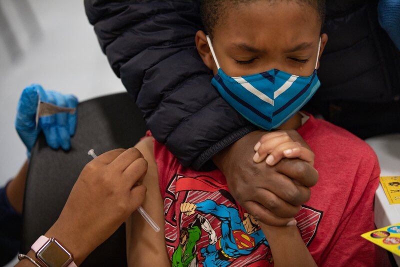 A child wearing a face mask and a red shirt holds an adult’s hand while getting a COVID-19 vaccine.