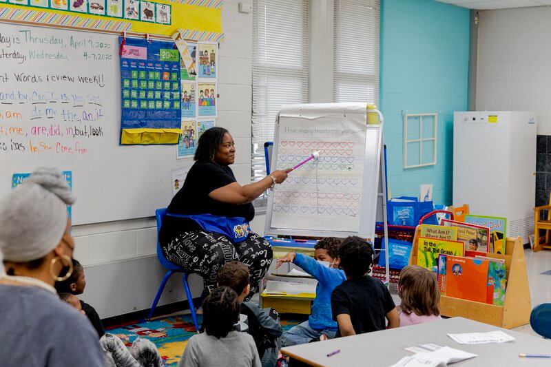 a teacher points to a white board while young children sit on a rug in front of her.