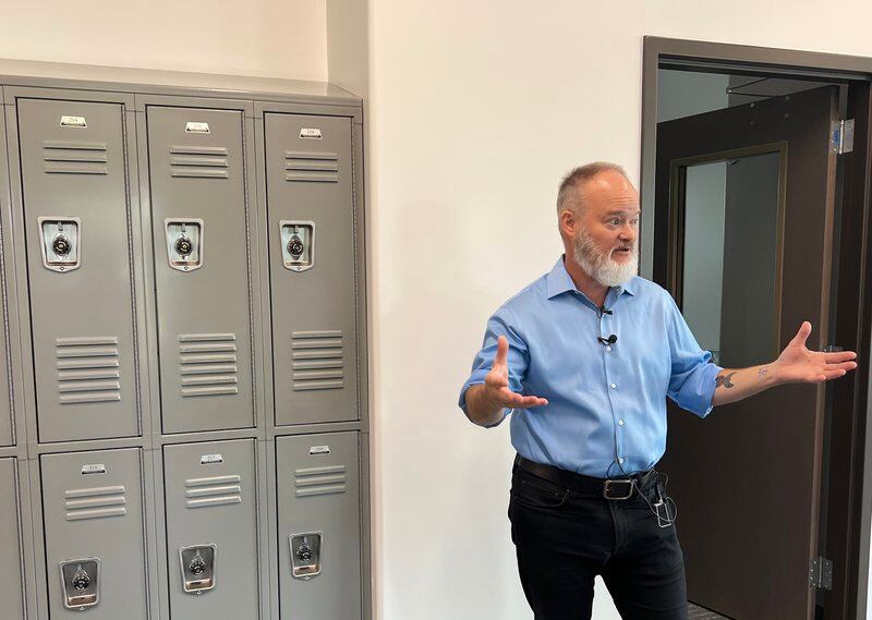 A man in a blue shirt holds out his hands while explaining the building with lockers to the left of him.