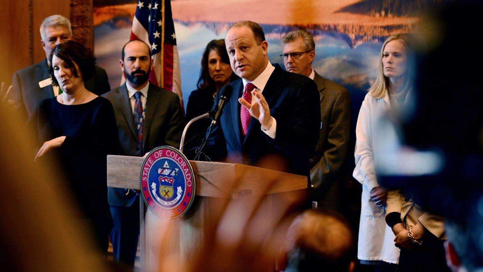 Colorado Governor Jared Polis speaks at a State of Colorado podium, surrounded by several people.