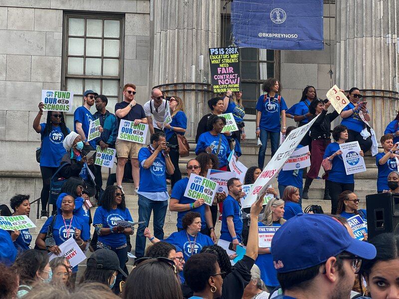 A group of people wearing bright blue T-shirts stand on the steps holding signs for a fair contract. 