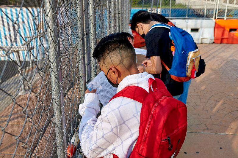 High school students complete their COVID screening forms next to a chainlink fence outside of their school’s entrance. 