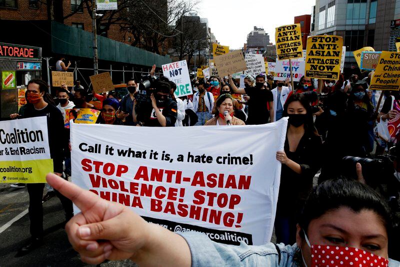 A crowd of people protest against Anti-Asian violence in New York City. A woman points in front of a banner that reads, “Call it what it is, a hate crime! Stop Anti-Asian Violence! Stop China Bashing!”