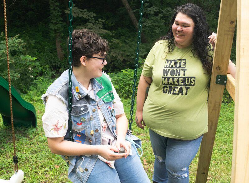 A youth in jeans and a jeans vest sits on a swing next to a person in a yellowish t-shirt that reads “Hate won’t make us great.”
