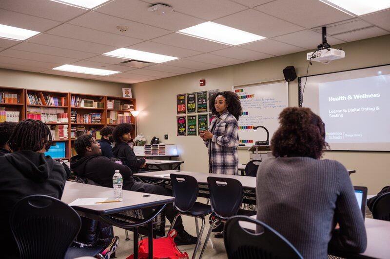A woman wearing a plaid top teaches a class to a group of high school students.