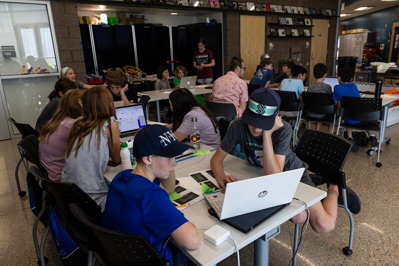 Teenagers work on laptops at tables in a crowded classroom. They’re working in small groups and sharing computers.