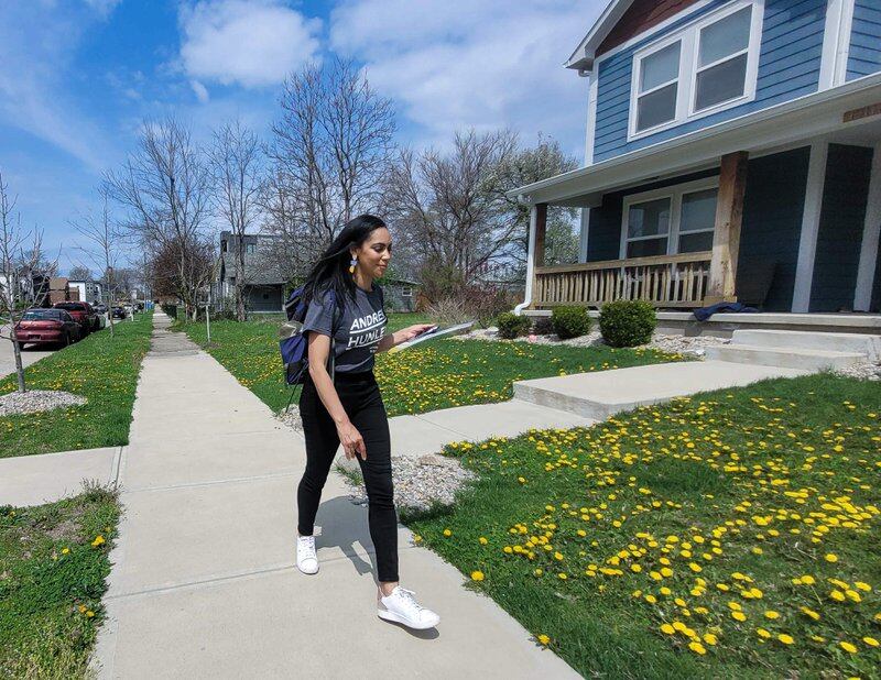 IPS principal Andrea Hunley, wearing a navy blue “Andrea Hunley” T-shirt, black stretch pants, and white athletic shoes, and carrying a clipboard and backpack, walks on a sidewalk past lawns in the Martindale-Brightwood as she campaign for state Senate.