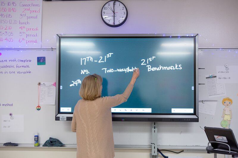 A female middle school teacher writes testing dates on a green SMART board.